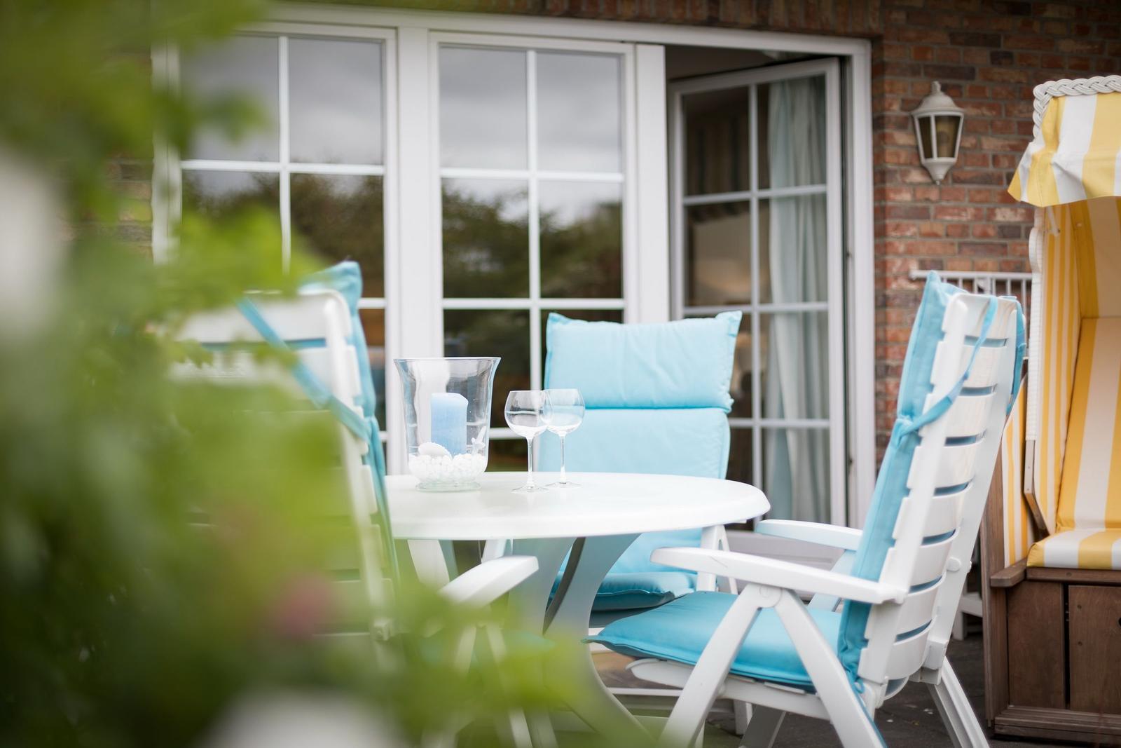 Terrasse mit weißem Tisch, blauen Sitzkissen und Glasgefäßen.