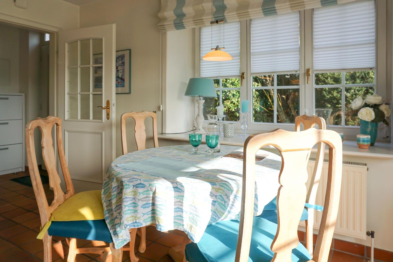 Runder Tisch mit Stühlen und Tischdecke mit Fischmotiv vor Fenster mit Jalousien.
