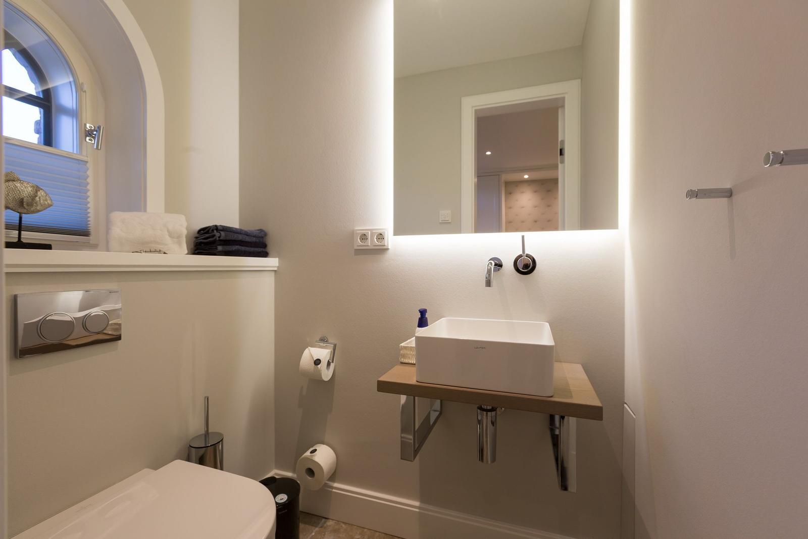 Badezimmer mit Waschbecken, Spiegel und Toilette. Fenster mit Jalousie.