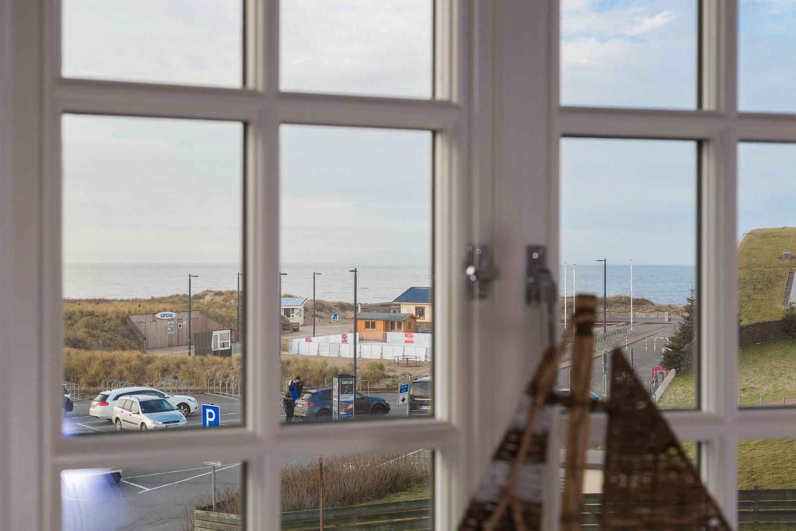 Durch das Fenster blickt man auf einen Strandparkplatz und das Meer.