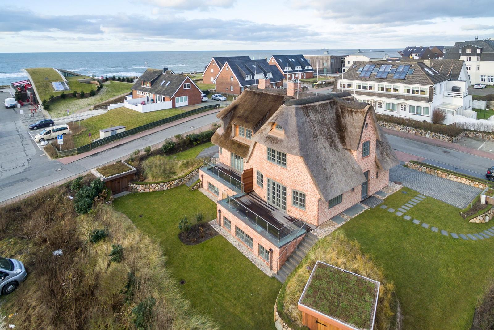 Haus mit strohgedecktem Dach und Glasbrüstung, umgeben von Grünflächen und Strand.
