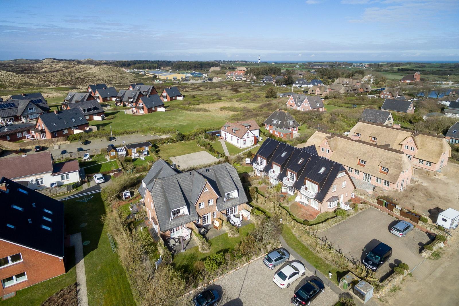 Aerial view of a village with houses, green fields, and a distant lighthouse.