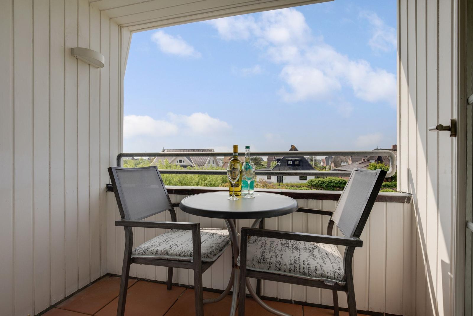 Balcony with table and two chairs. View of rooftops and sky.