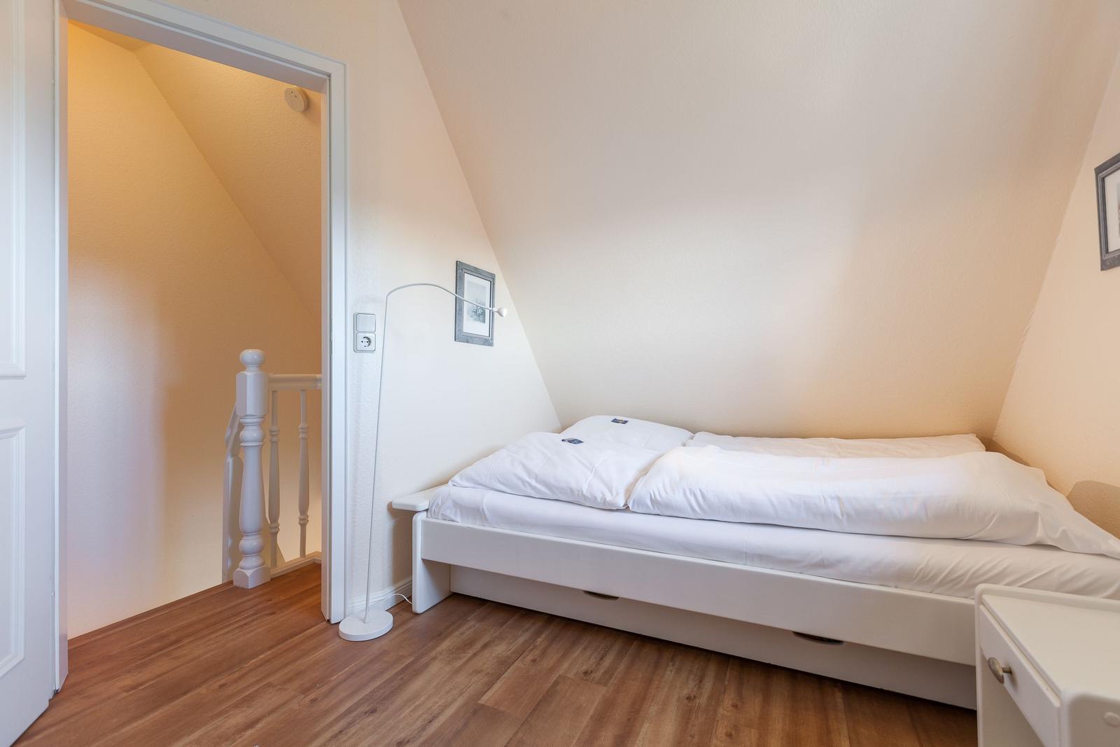 Schlafzimmer mit weißem Bett, Holzboden und schrägen Wänden. Tür führt zu Treppenhaus.