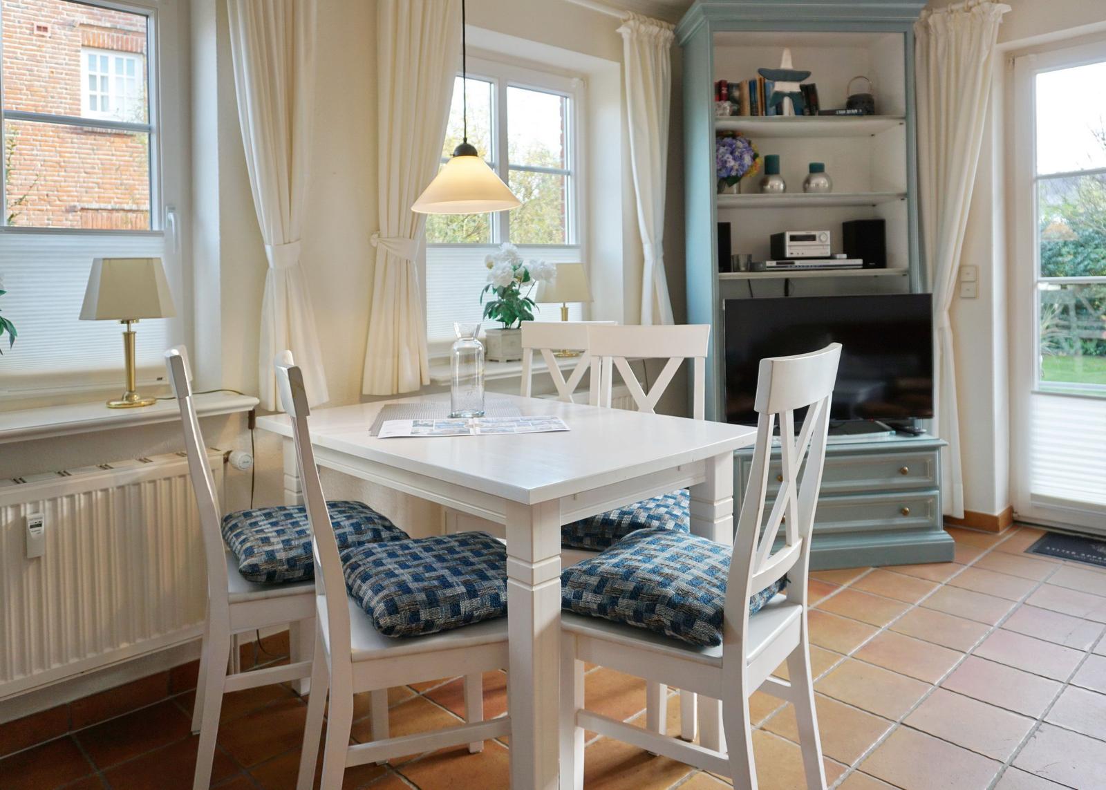 Essbereich mit weißem Tisch, Stühlen und blauen Kissen. Fernseher und Bücherregal im Hintergrund.