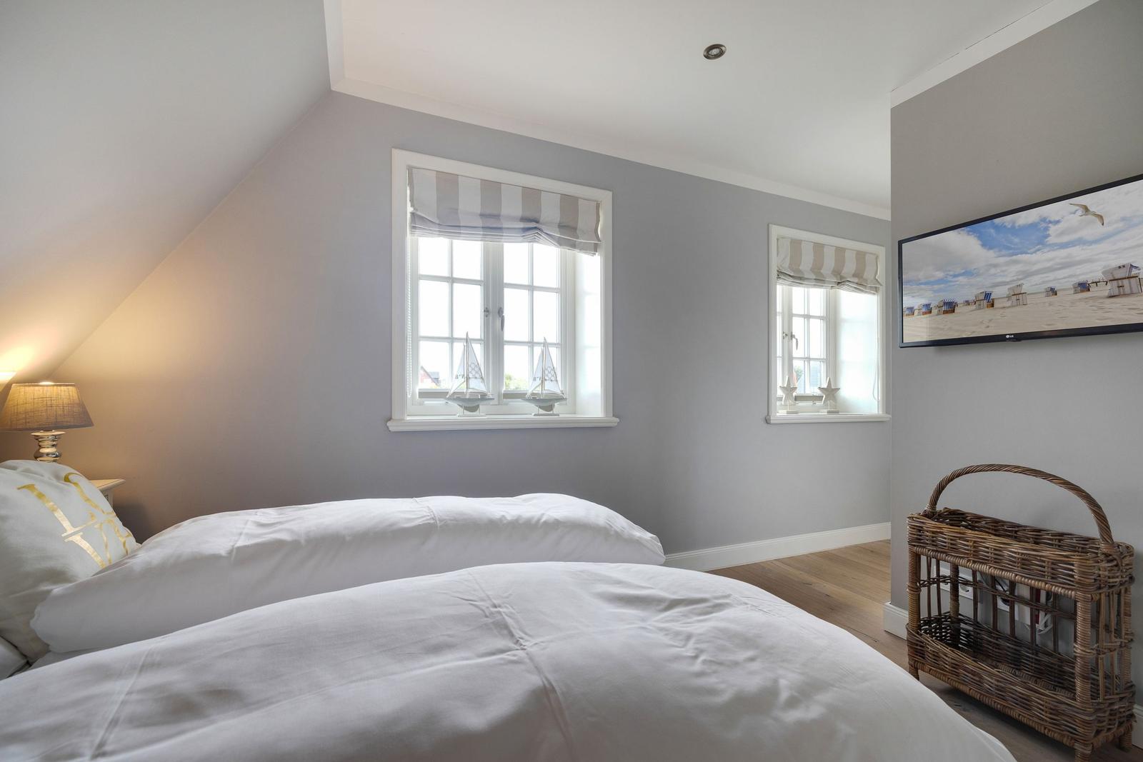 Schlafzimmer mit Bett, zwei Fenstern und Fernseher an der Wand.