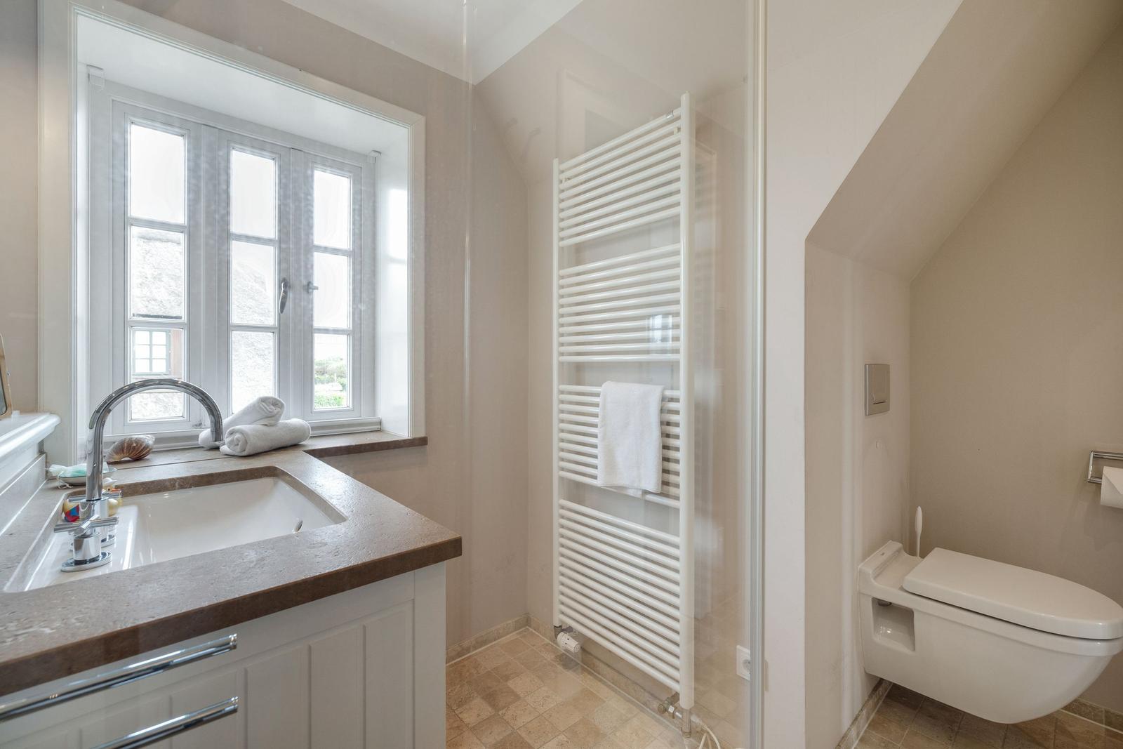 Badezimmer mit Waschbecken, Fenster und Heizkörper. Toilette im Anschluss.