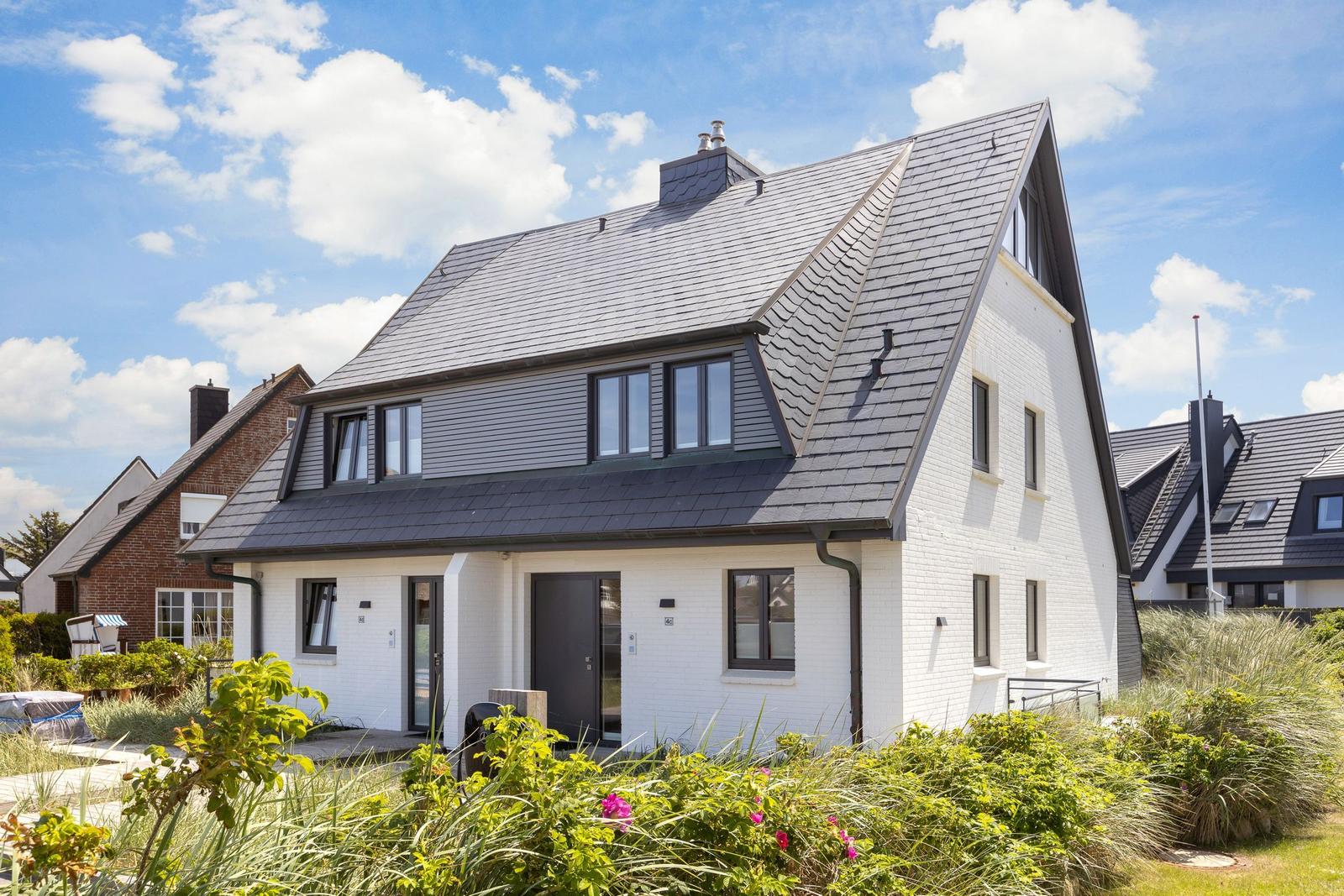Modern villa with white walls and dark roof. Garden with plants and flowering blooms.