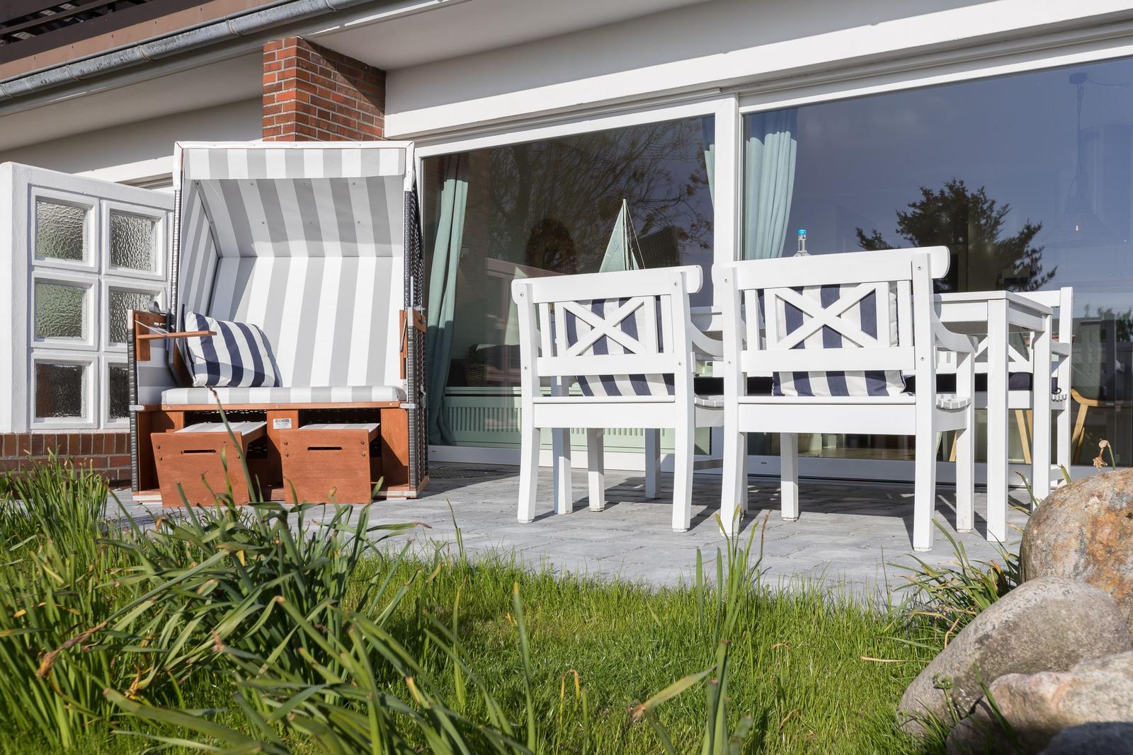 Terrace with beach chair and white chairs. Glass facade and greenery in the background.