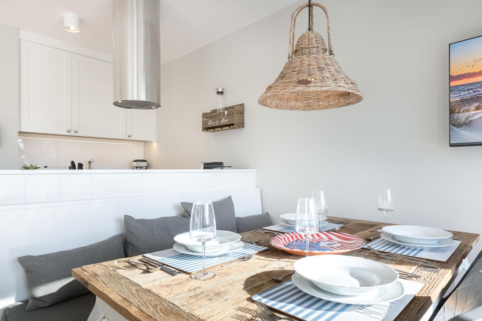 Dining area with rustic wooden table and gray bench. Hanging lamp and kitchen area in background.