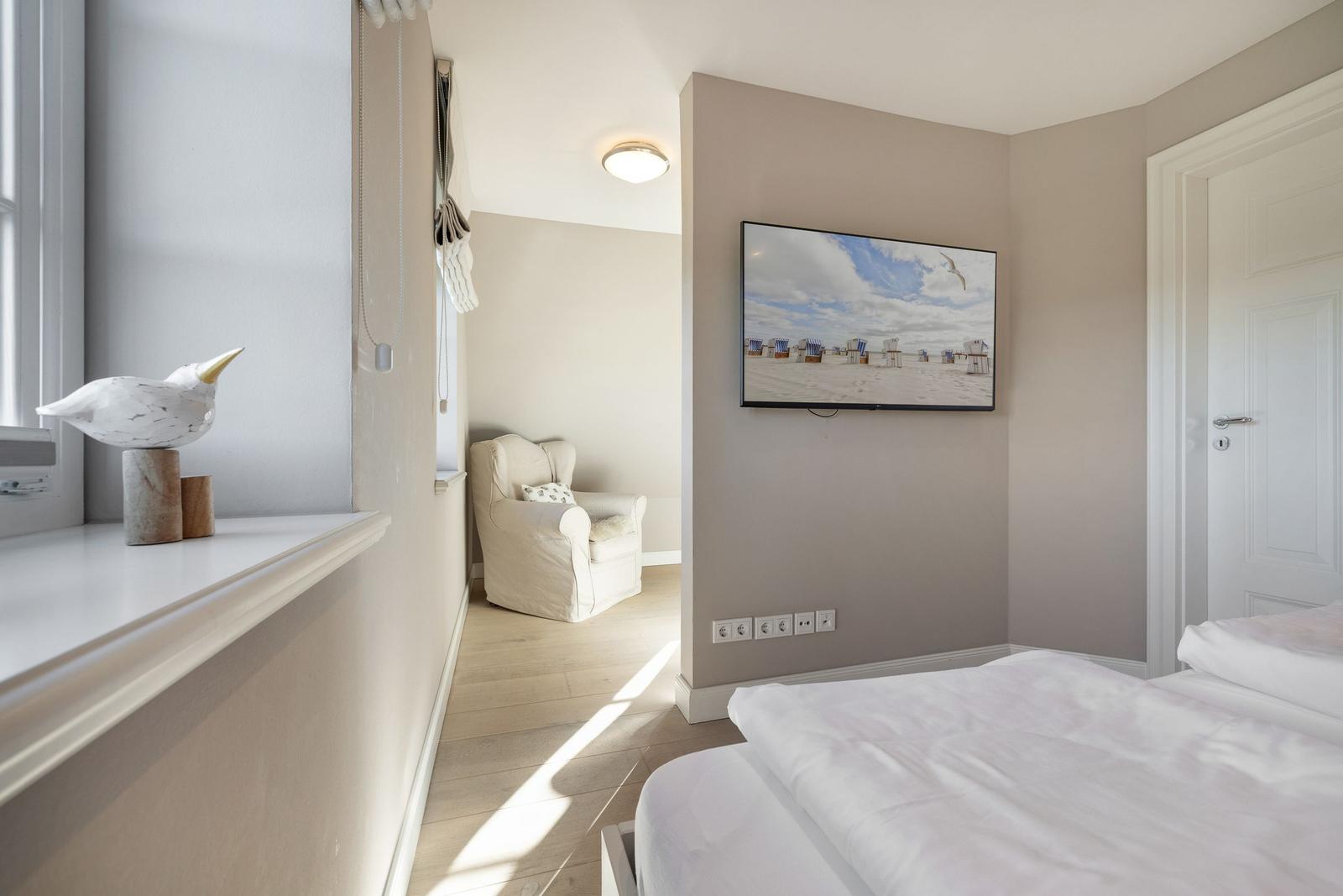 Schlafzimmer mit Bett, Fernseher und Sitzgelegenheit. Fensterbank mit Vogeldekoration.