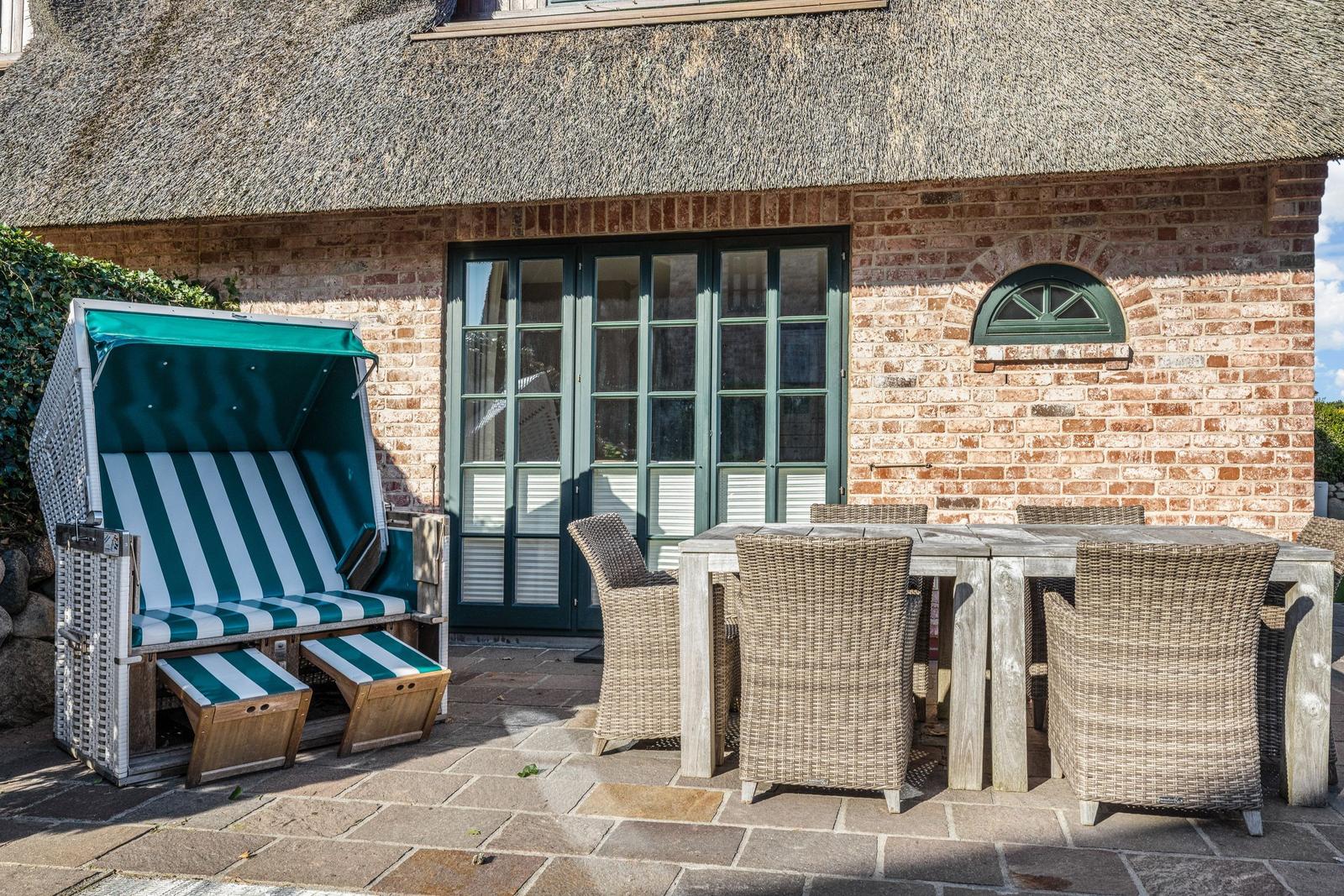 Terrasse mit Strandkorb und Esstisch vor einem Haus mit Strohdach und Backsteinmauer.