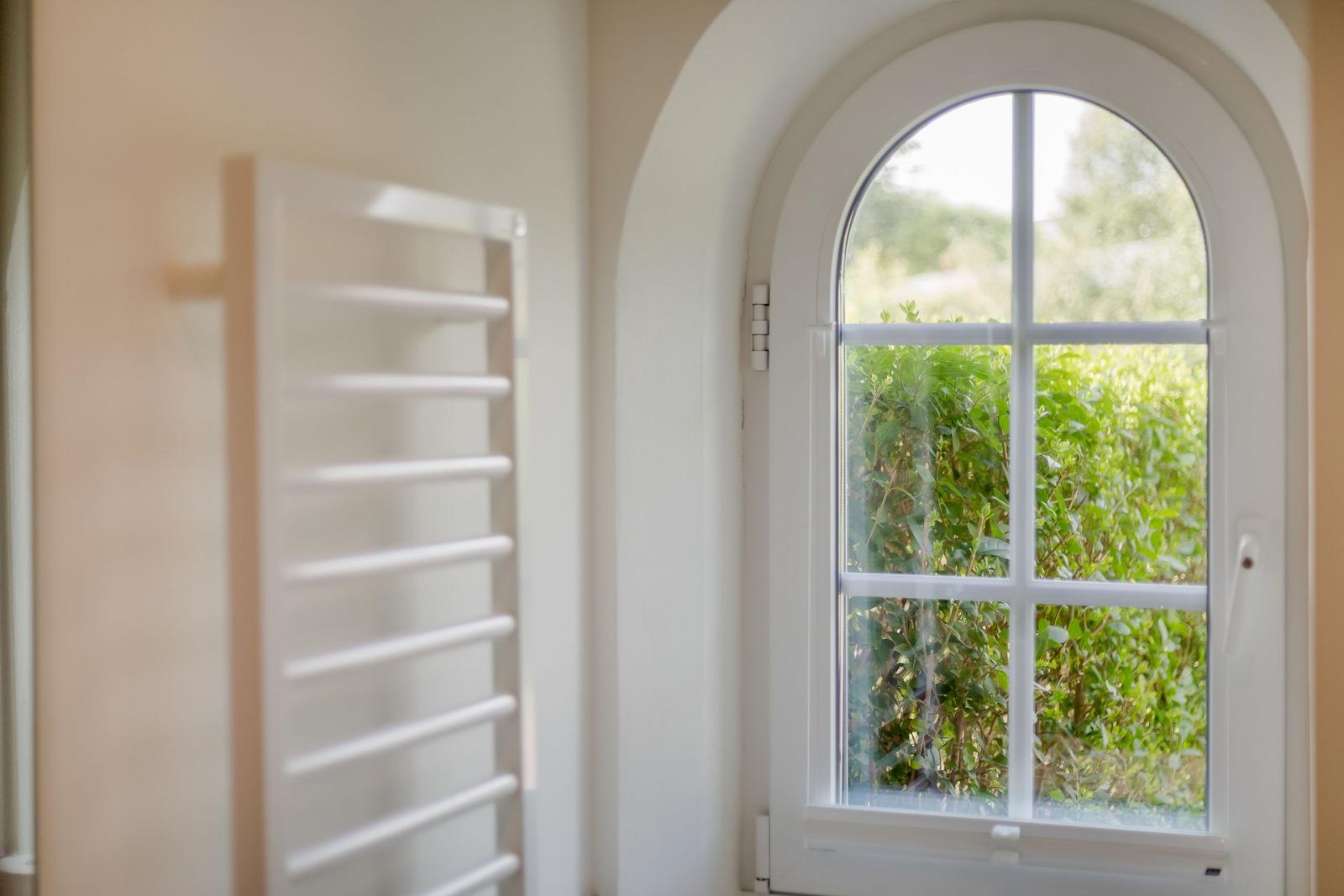 Badezimmer mit Heizkörper und Bogenfenster mit Blick auf grüne Hecke.