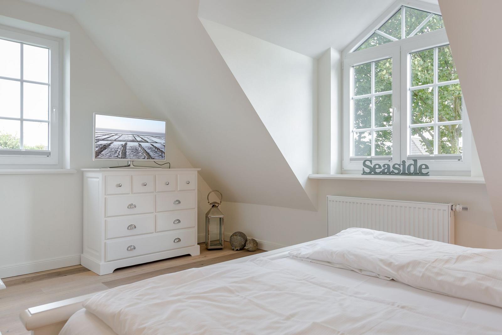Schlafzimmer mit Dachschrägen, weißem Bett und TV-Schrank. Fenster mit Blick auf Bäume.