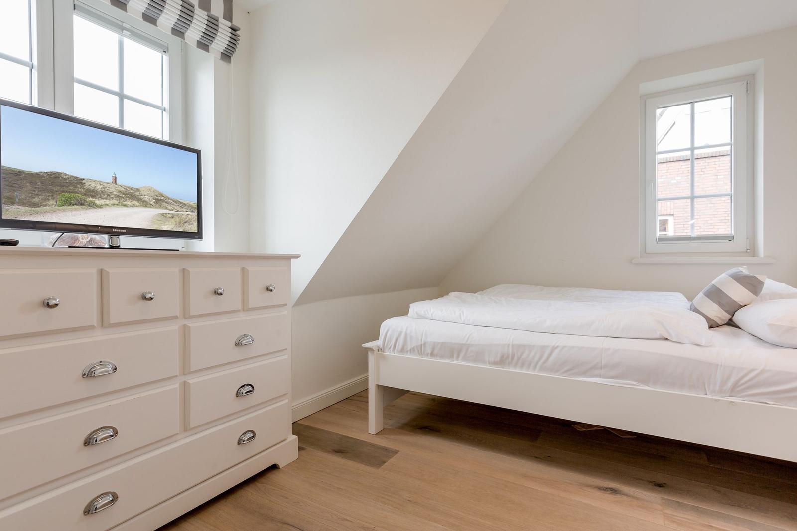 Schlafzimmer mit weißem Bett, Schrank und Fernseher. Holzboden und schräge Wand.
