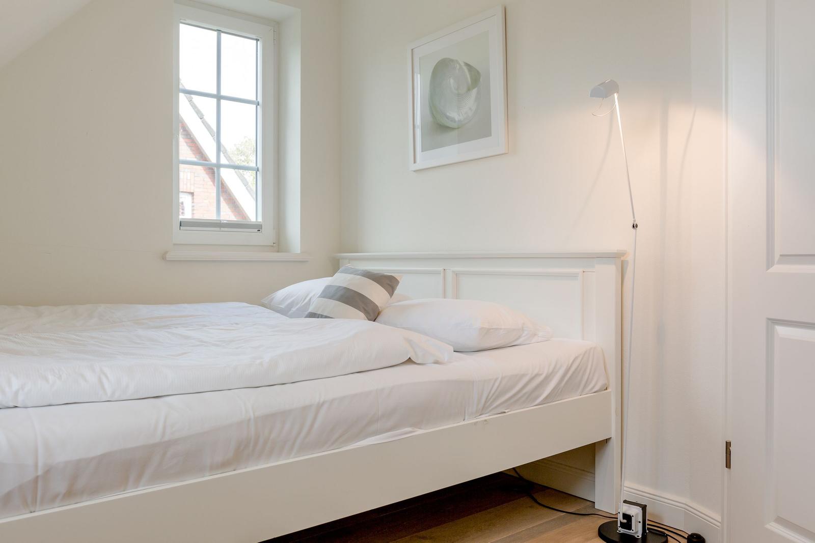 Ein Bett mit weißer Bettwäsche, Wandbild und Stehlampe neben einem Fenster.