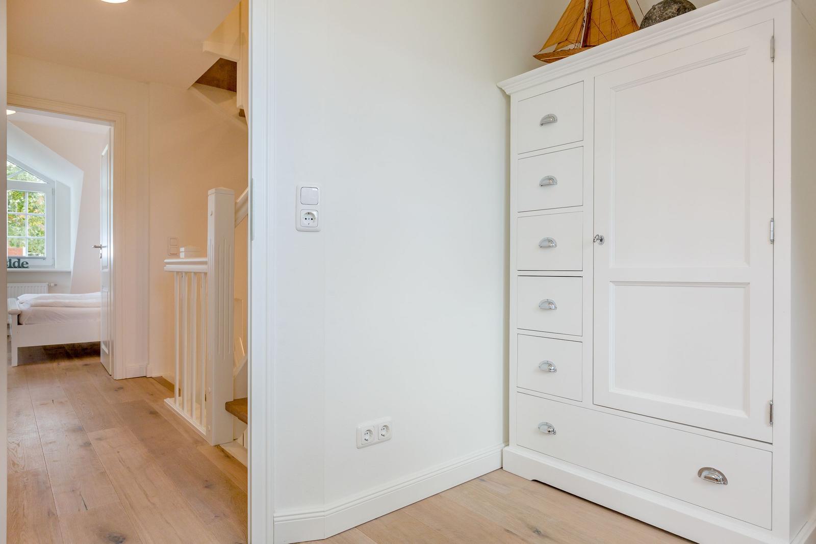 Weißer Schrank mit Schubladen und Tür. Blick in Schlafzimmer mit Bett und Fenster.