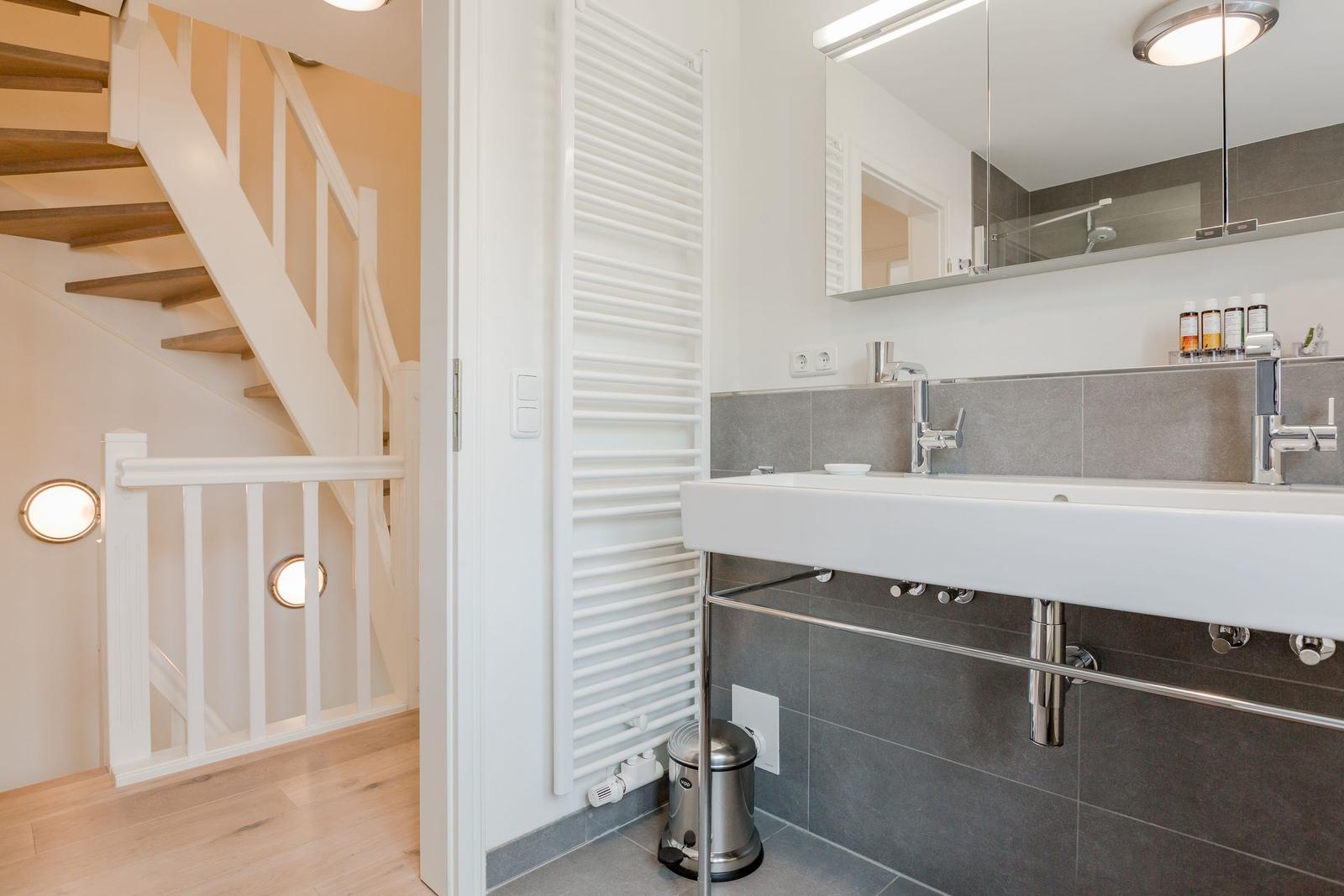 Badezimmer mit Waschbecken, Spiegel und Heizkörper. Treppenhaus im Hintergrund.