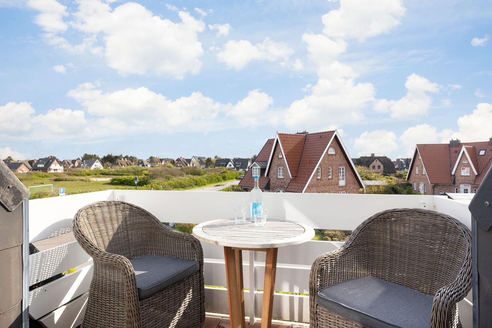 Terrace with table and two chairs. View of houses and green areas under blue sky.