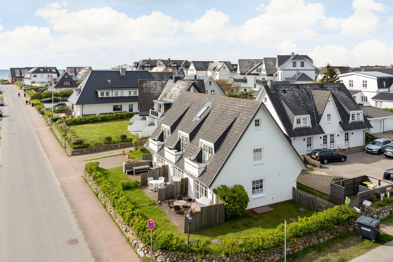 White house with gray roof and garden. Street with cars and view of the sea.