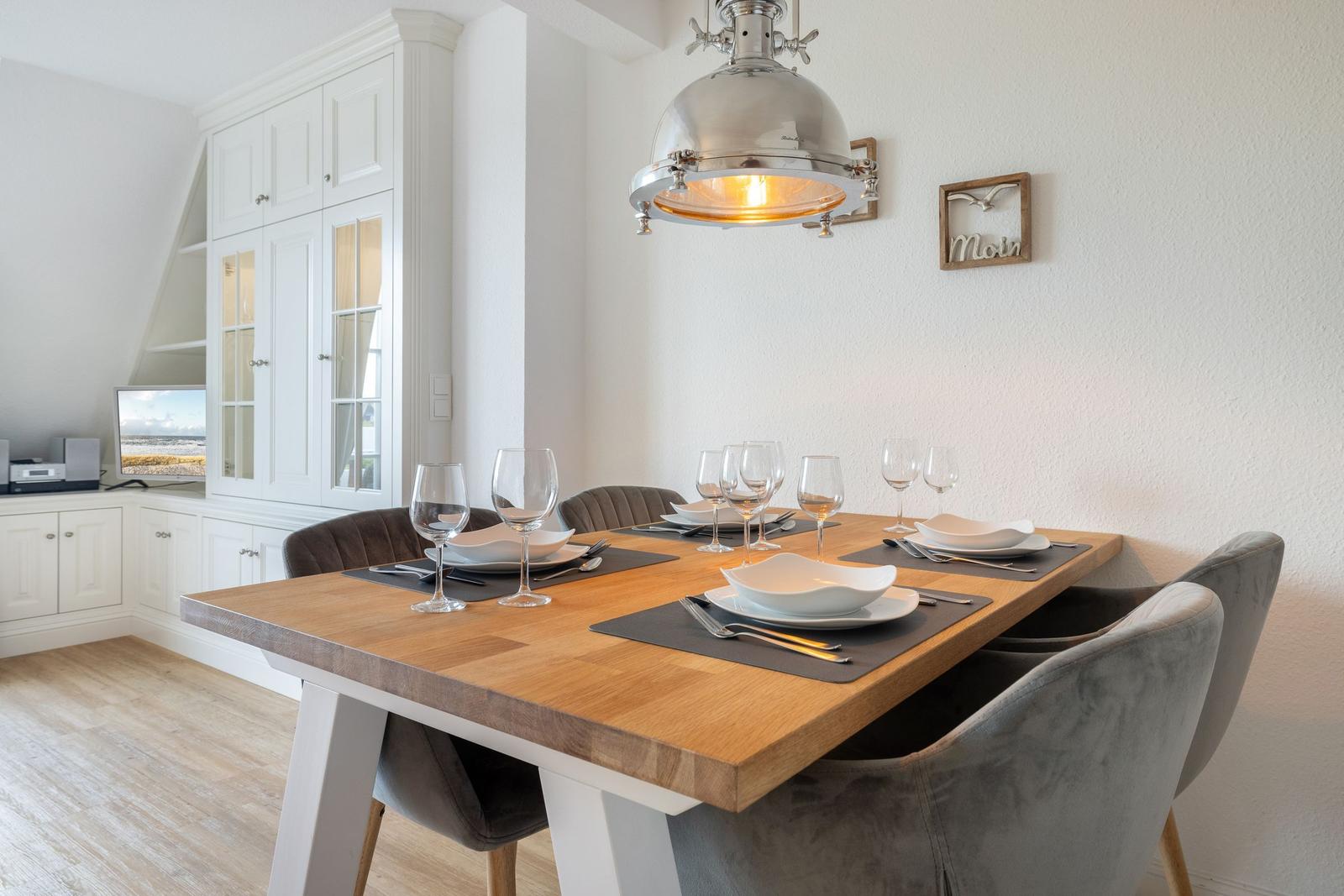Dining room with wooden table, chairs, and pendant light. Kitchen cabinets in background.
