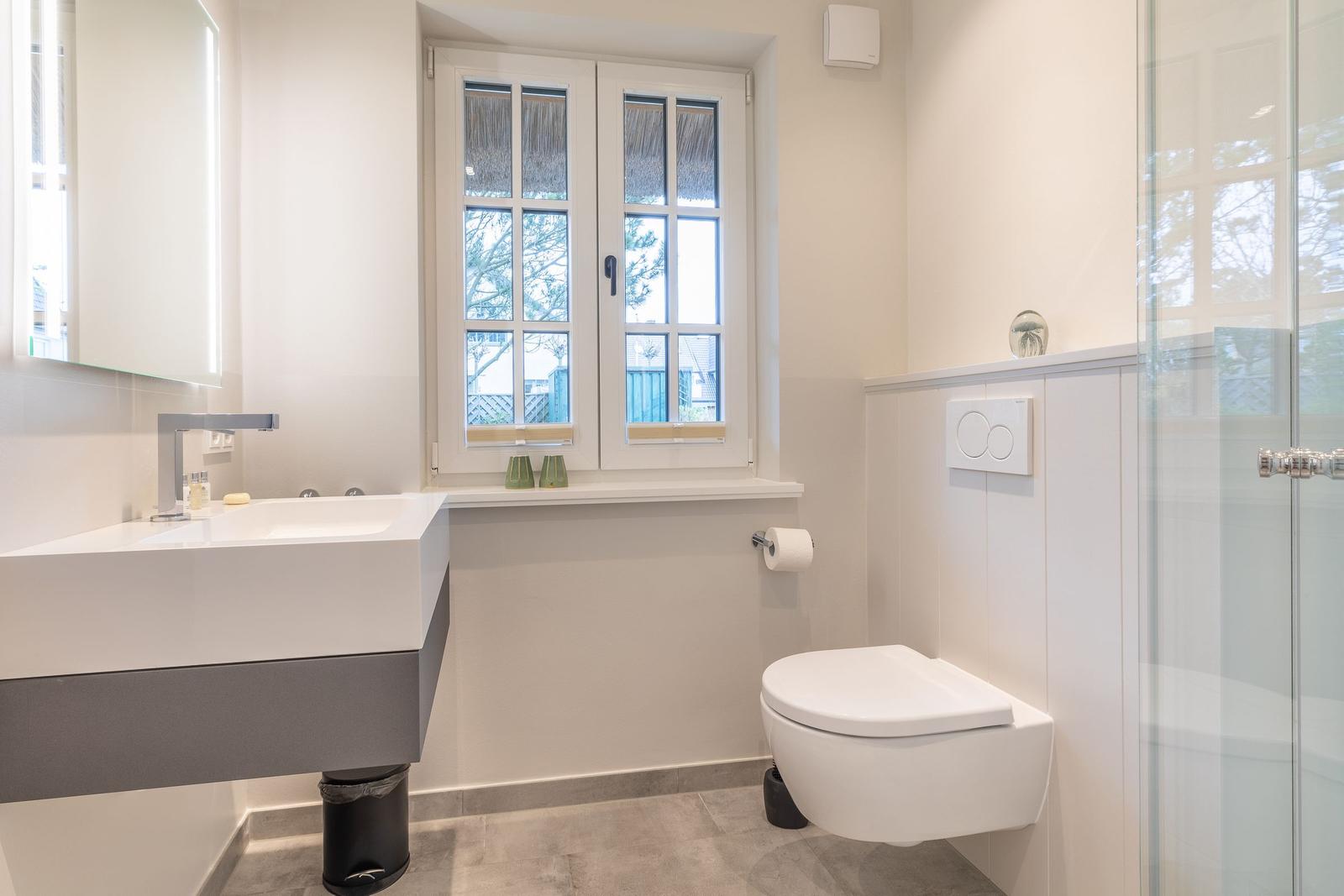 Badezimmer mit Waschbecken, Toilette und Dusche. Fenster mit Blick nach außen.