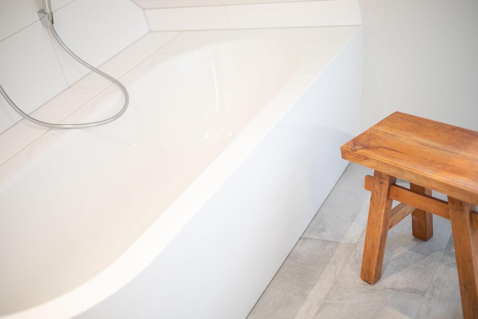 Eine weiße Badewanne mit Duschkopf und hölzerner Bank.