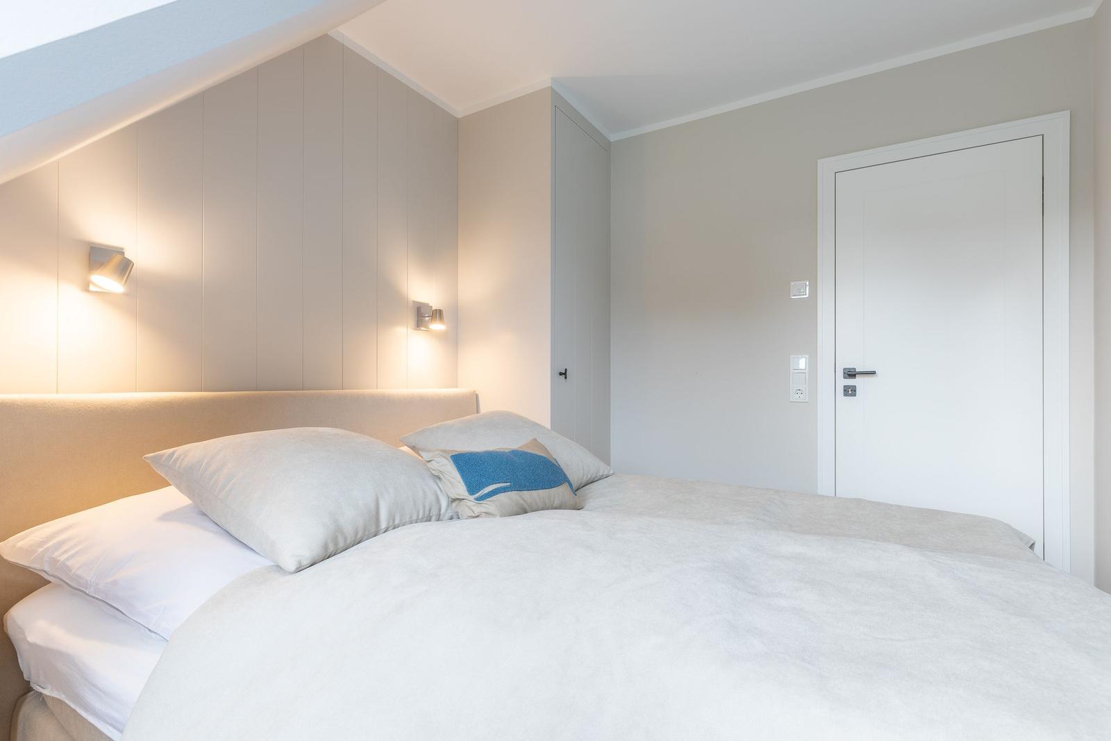 Schlafzimmer mit Bett, Kopfteil und Wandlampen. Tür und Schrank sind sichtbar.