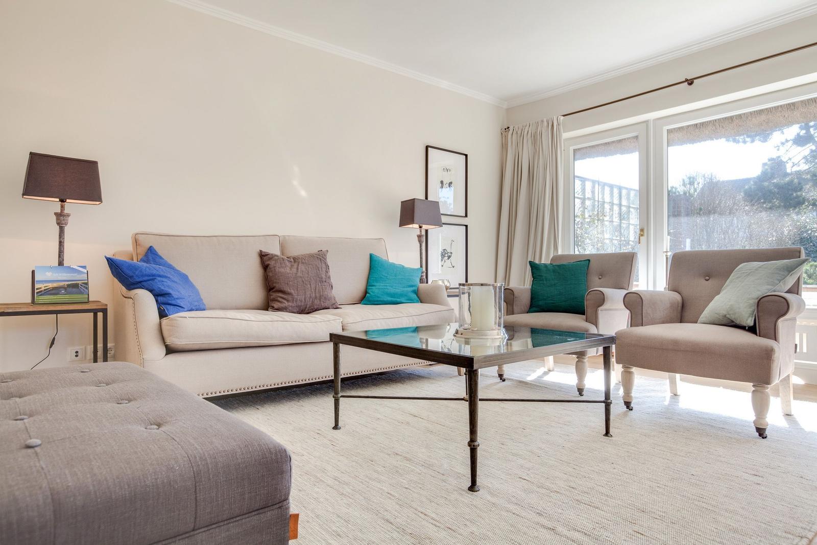 Living room with sofa, two armchairs, and glass table. Windows with curtains. Rug and lighting.