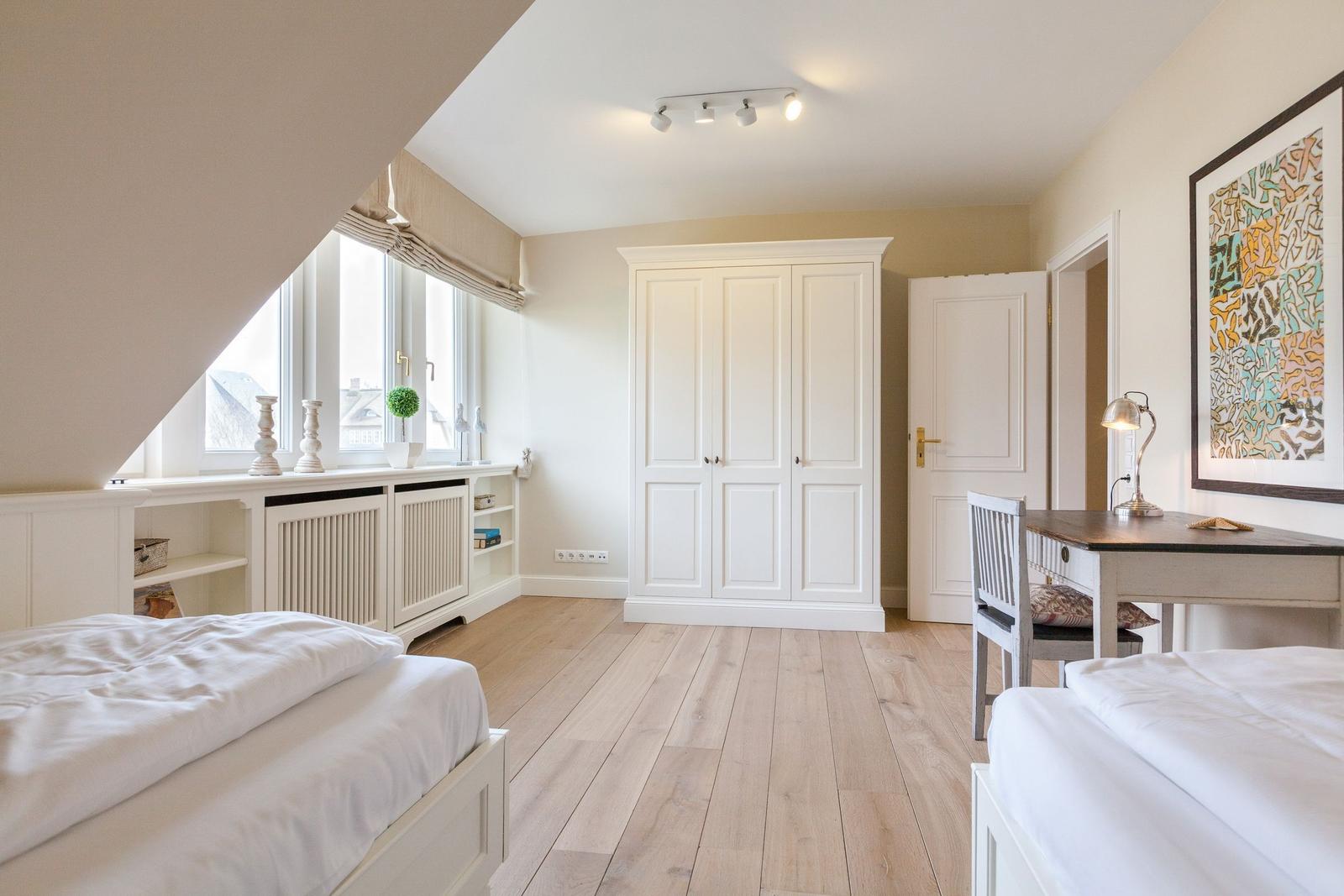 Room with two beds, desk, and wardrobe. Wooden floor and large window.