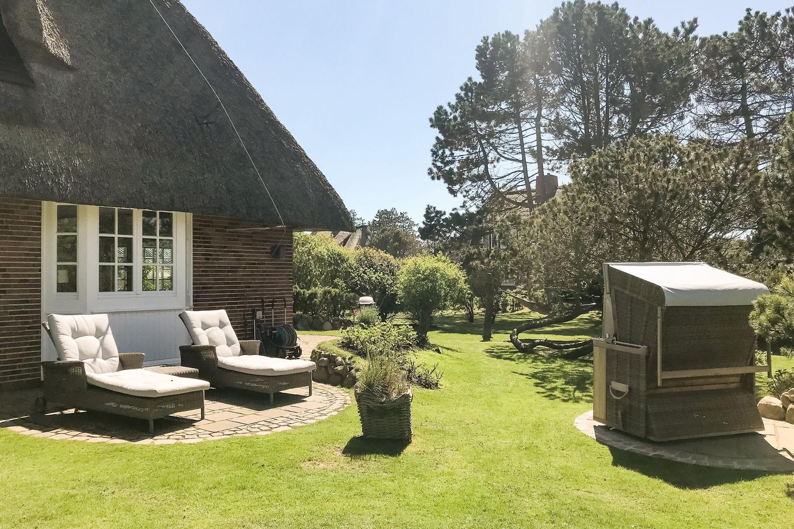 Backyard with loungers, lawn, and trees beside a thatched-roof house.