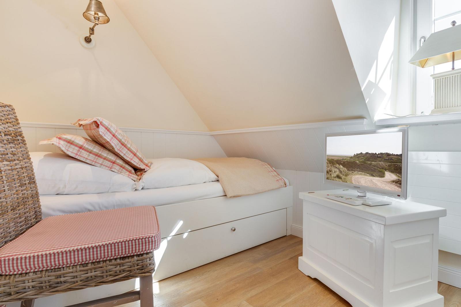 Schlafzimmer mit Dachschrägen, Bett, Wickerstuhl und Fernseher auf weißen Möbeln.