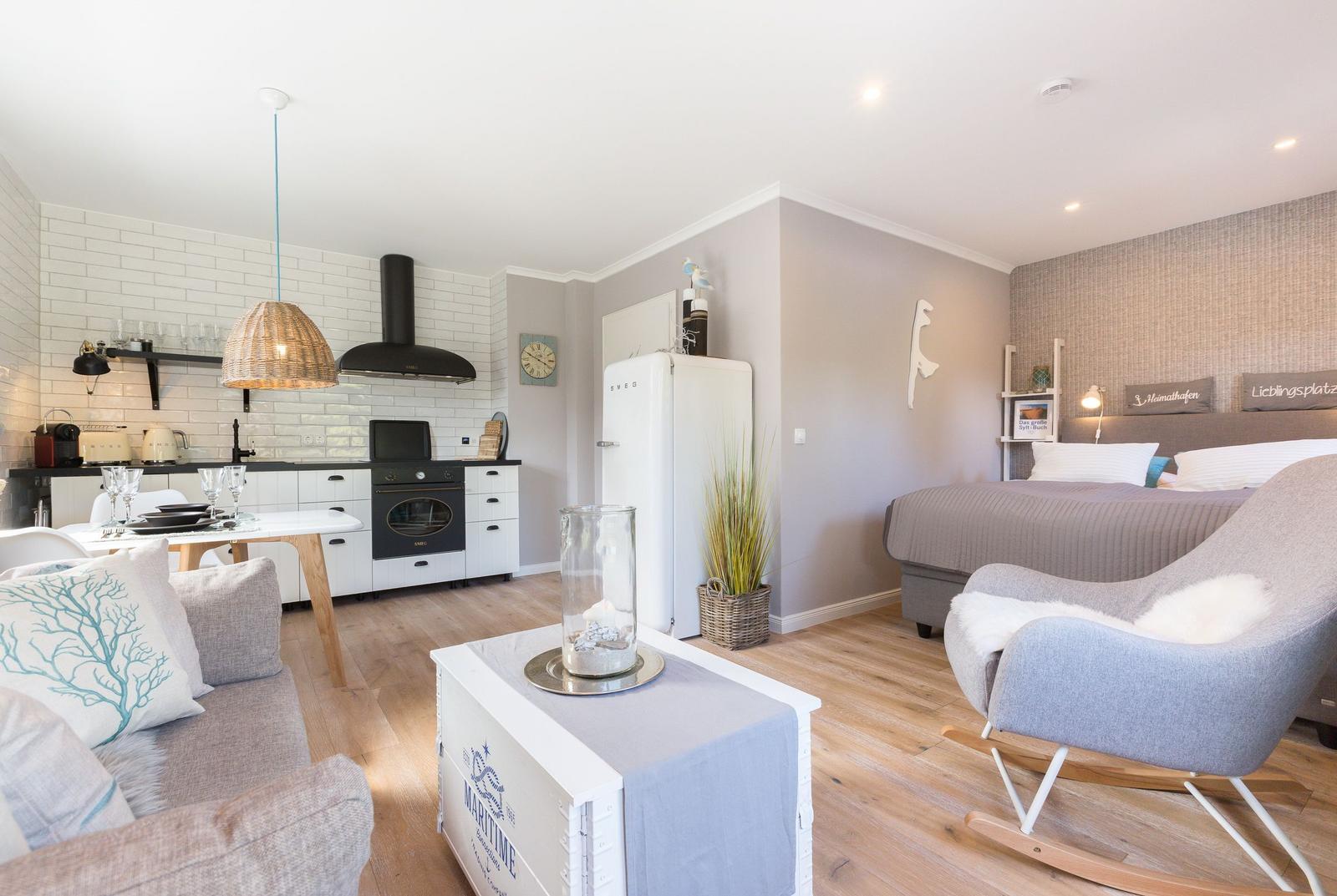 Living area with kitchen and bedroom. Wooden floor, white refrigerator, and bed with headboard.