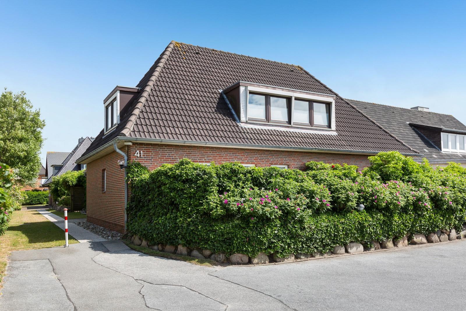 A house with brick facade and dormer windows, surrounded by green bushes with pink flowers.