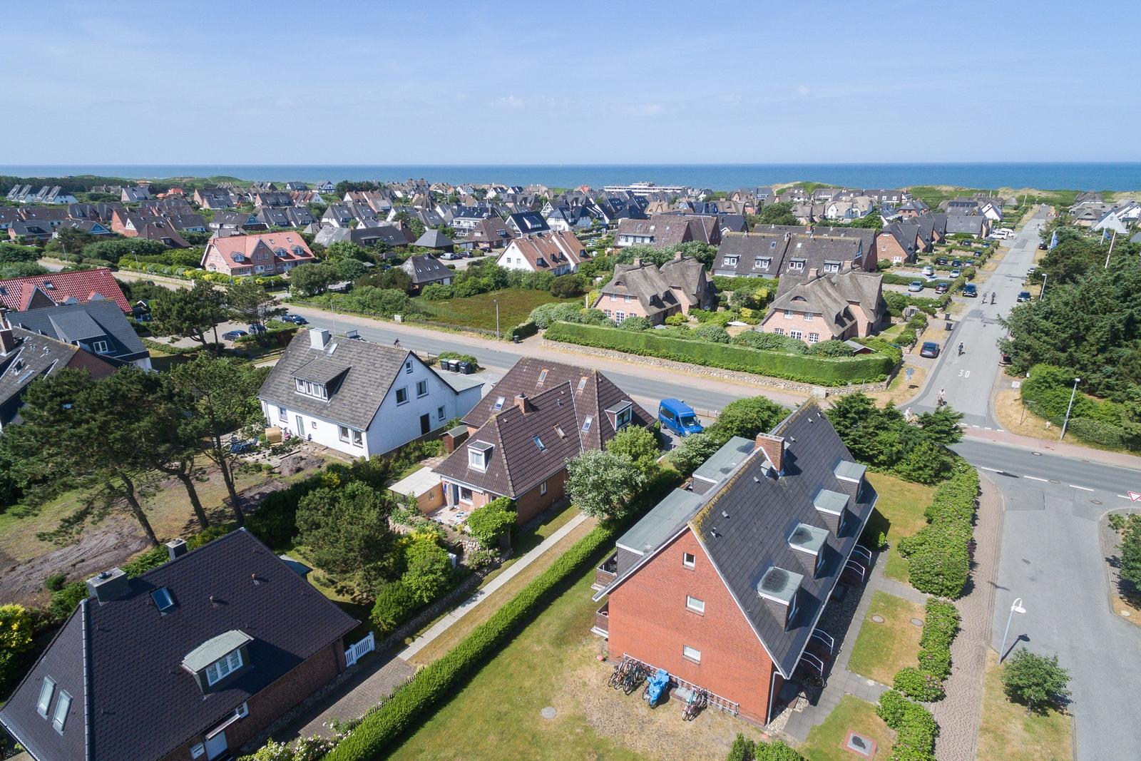 Aerial view of a coastal residential area with houses, greenery, and the sea in the background.