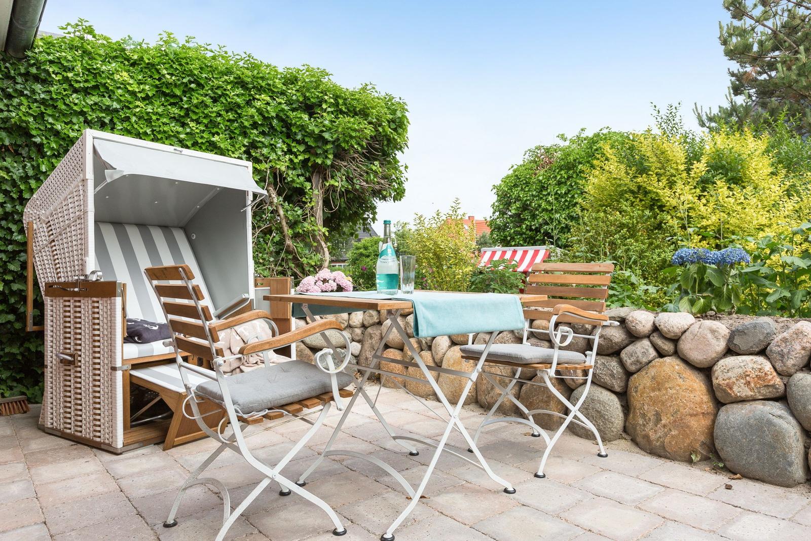 Terrace with lounge chair, table and chairs. Background: stone wall and green plants.