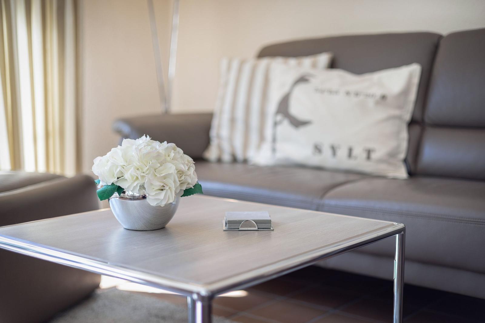Living room with gray sofa, table, and flowers. Pillow with Sylt logo.