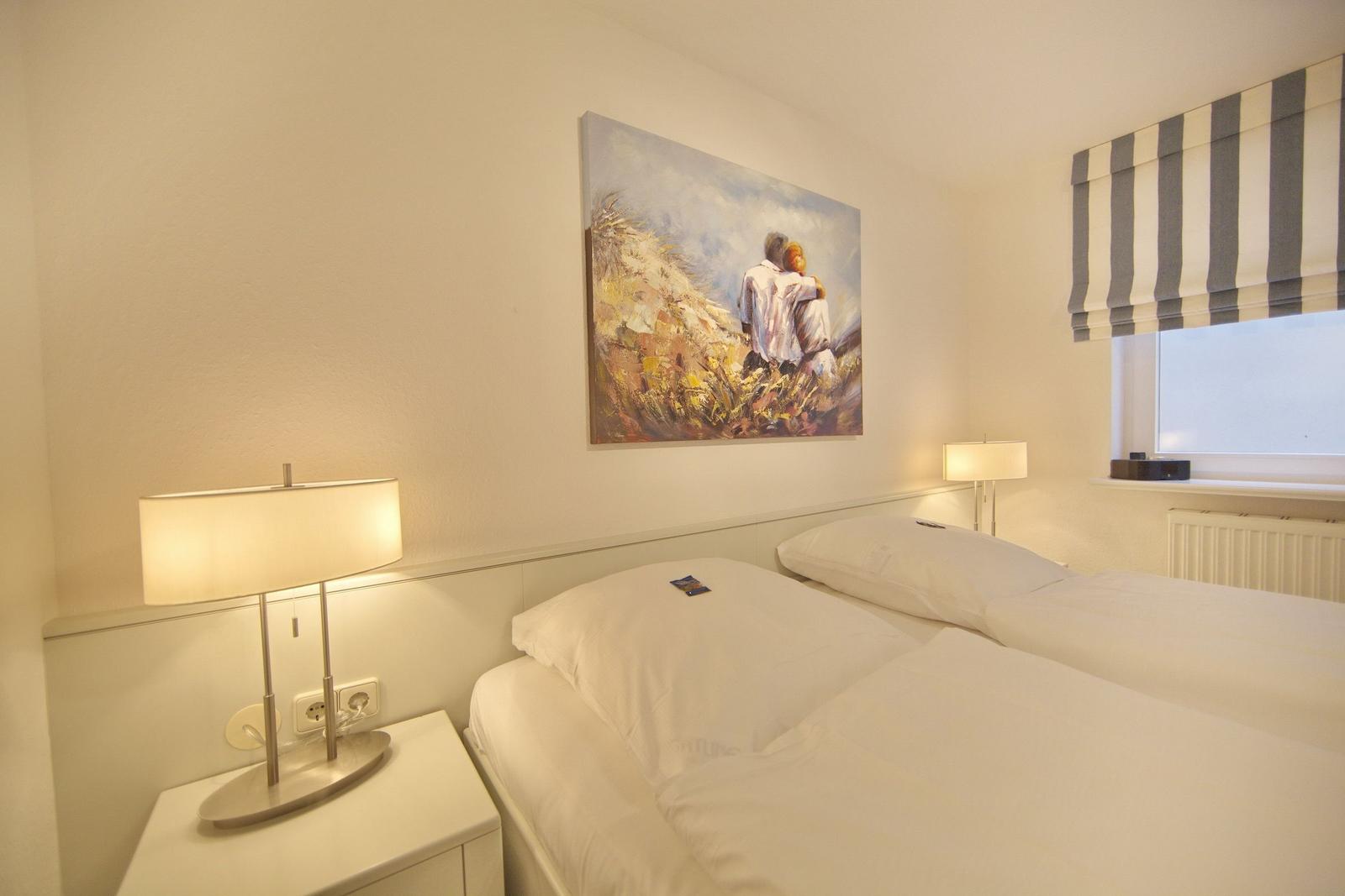 Two beds with white sheets, wall painting, and window with striped roller blind.