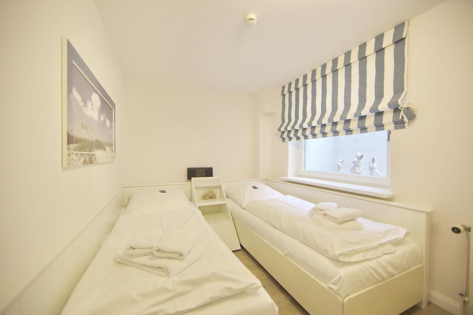Two beds with white linens and towels. Window with striped roller blind and white rabbit figurines.