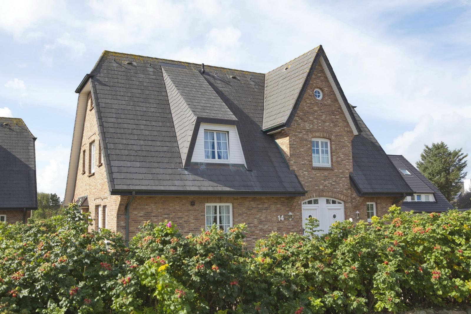 House with brick facade, dark roof, and white windows behind flower hedge.