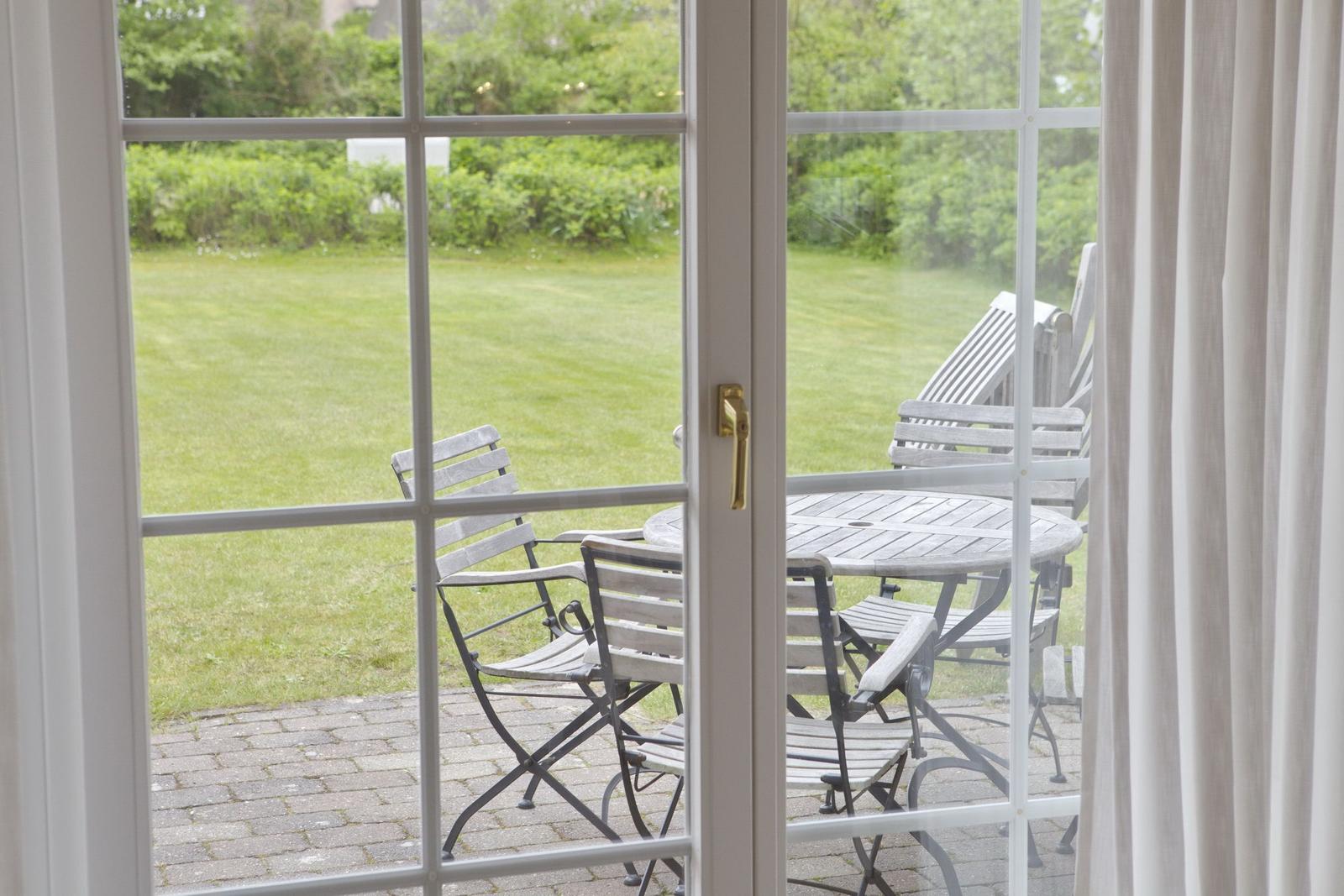 Through the glass door, a paved balcony with table and chairs is visible. Behind it lies a green lawn.
