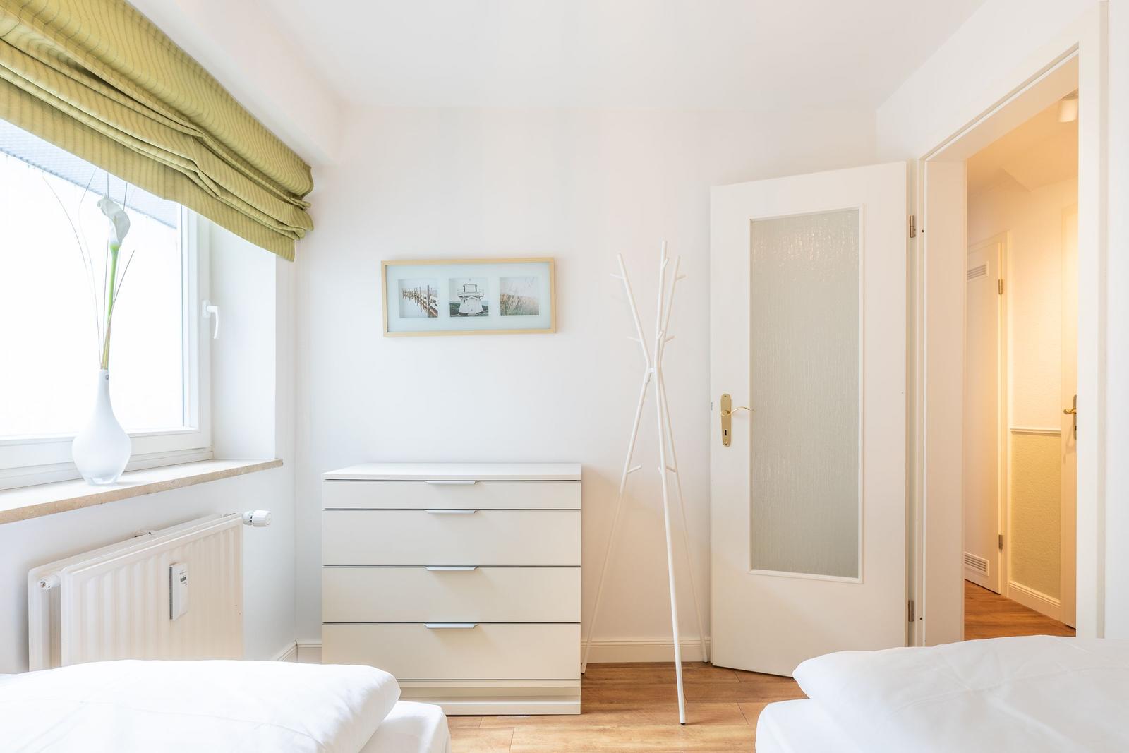 Schlafzimmer mit weißem Schrank, Fenster mit grünem Rolladen und offener Tür.