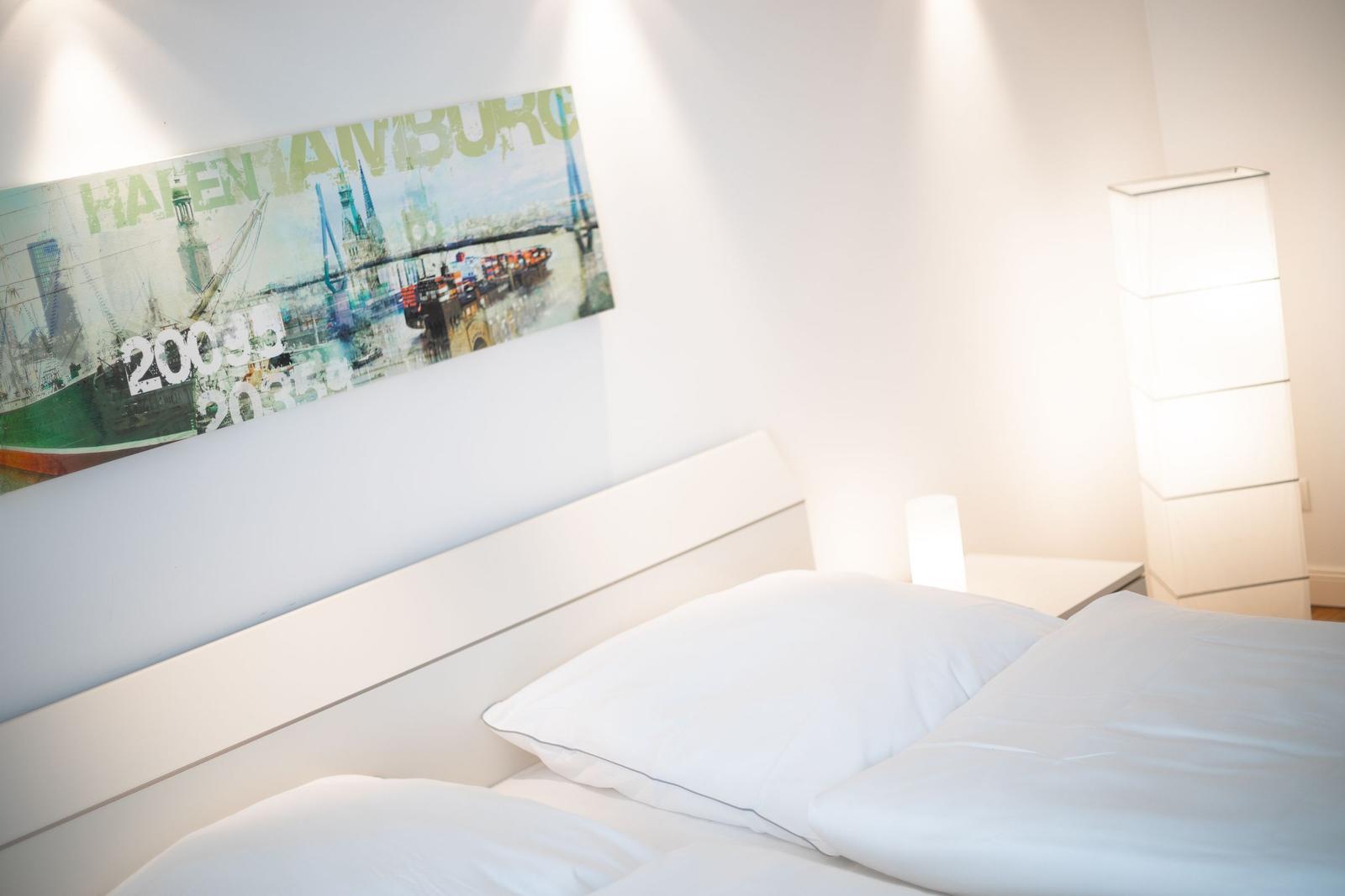 Schlafzimmer mit weißem Bett, Wandbild und Stehlampe.