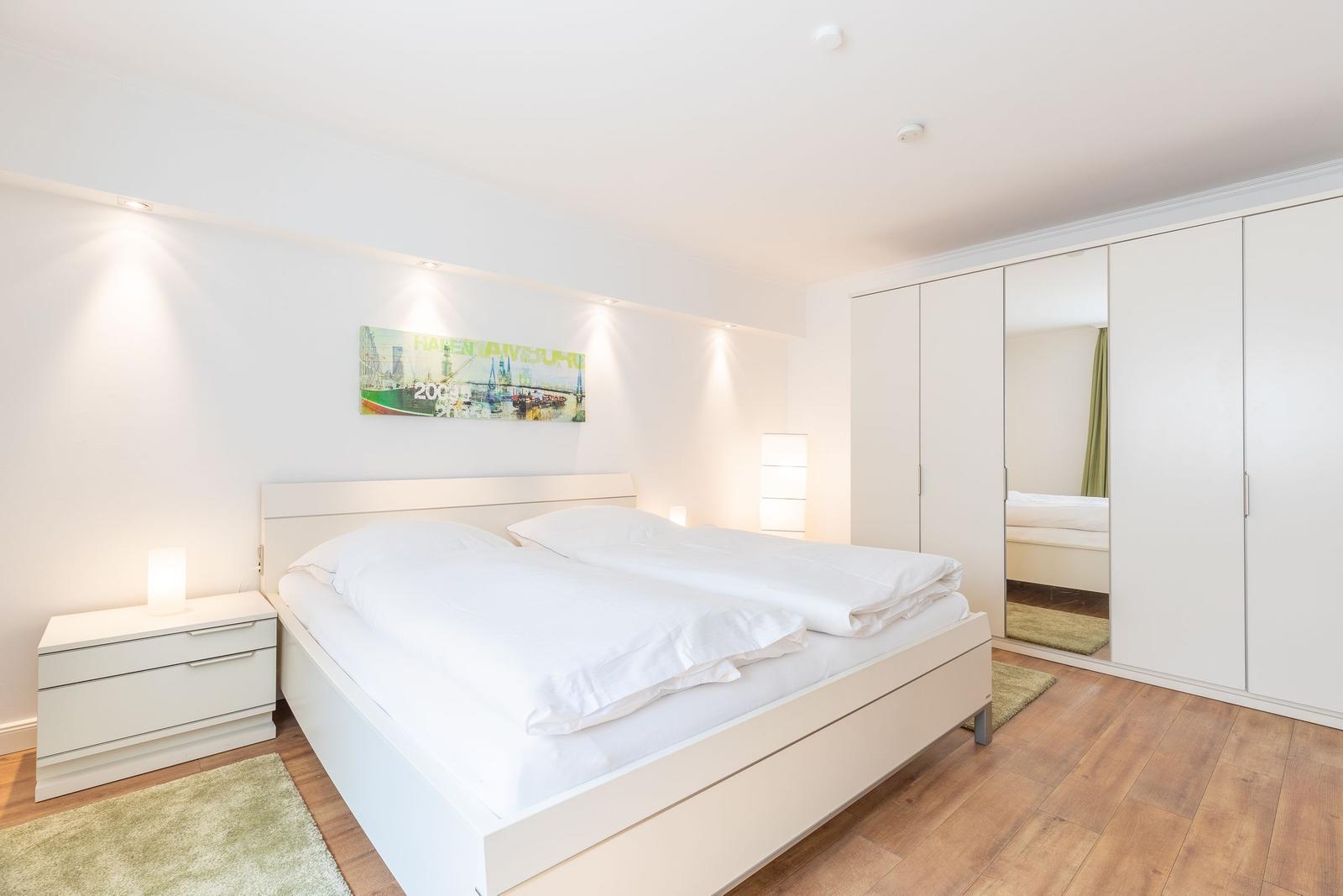 Schlafzimmer mit weißem Bett, Nachttisch und Kleiderschrank. Holzboden und Wandbild.