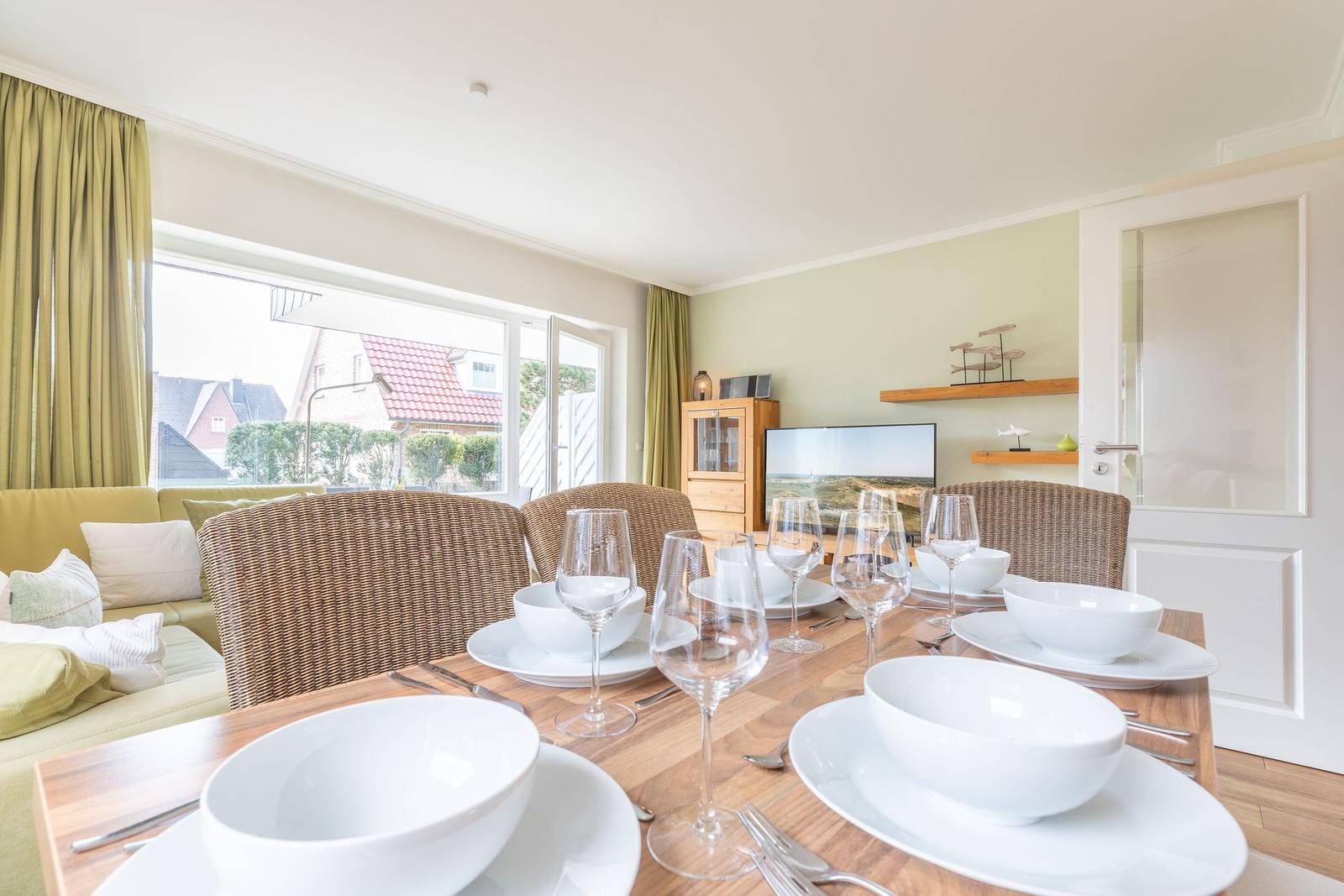 Esstisch mit weißen Tellern und Gläsern. Im Hintergrund Wohnbereich mit Sofa und TV.