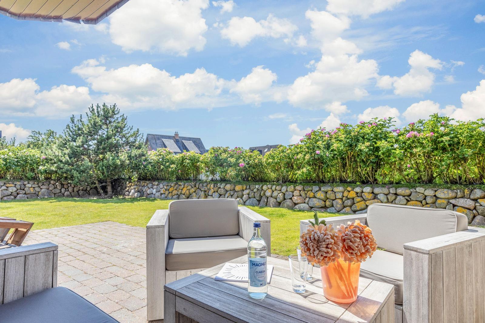 Terrace with seating, table, and flowers. Background: lawn, stone wall, and trees.