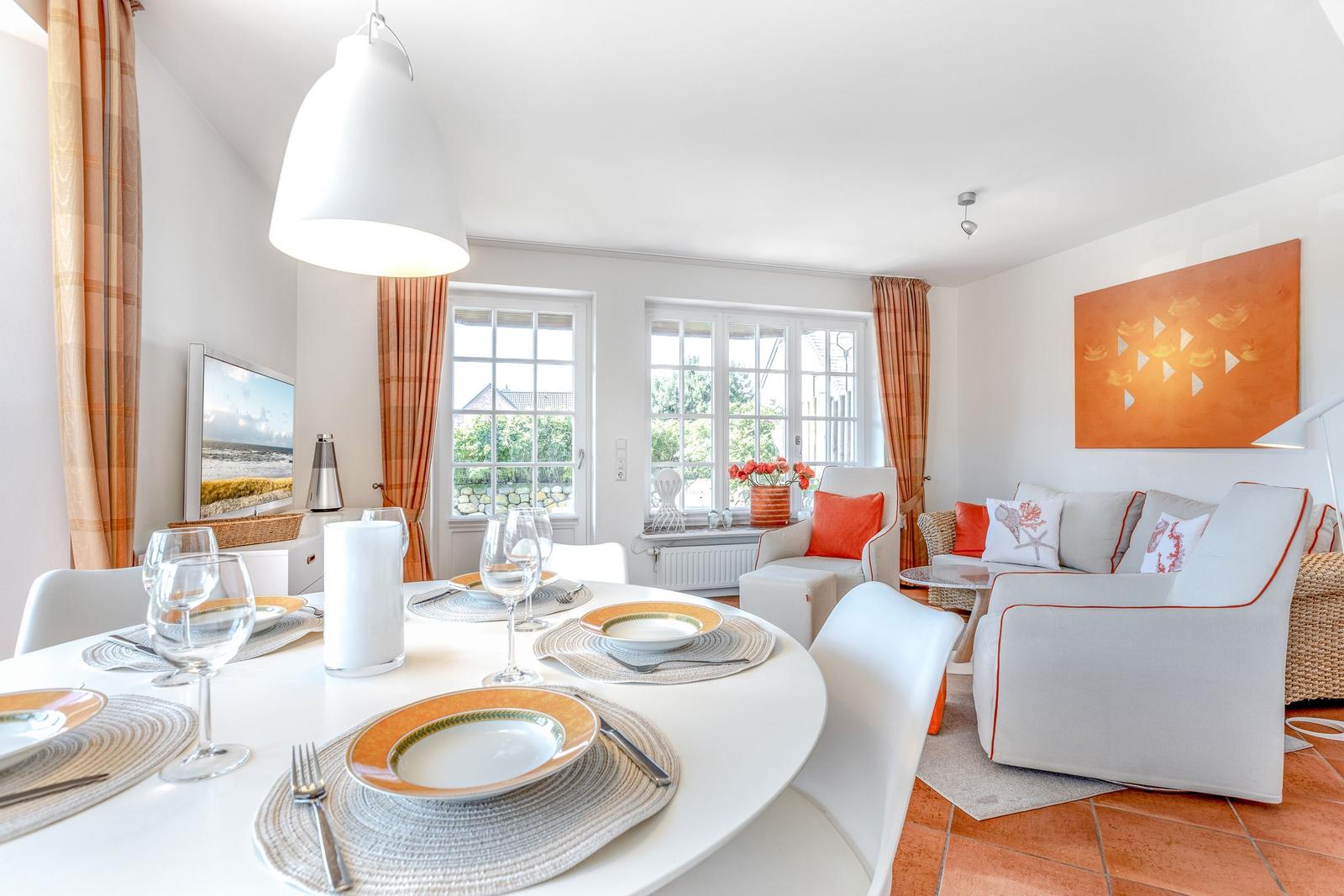 Dining room with round table, white chairs, and orange curtains. Living area with sofa and wall art.