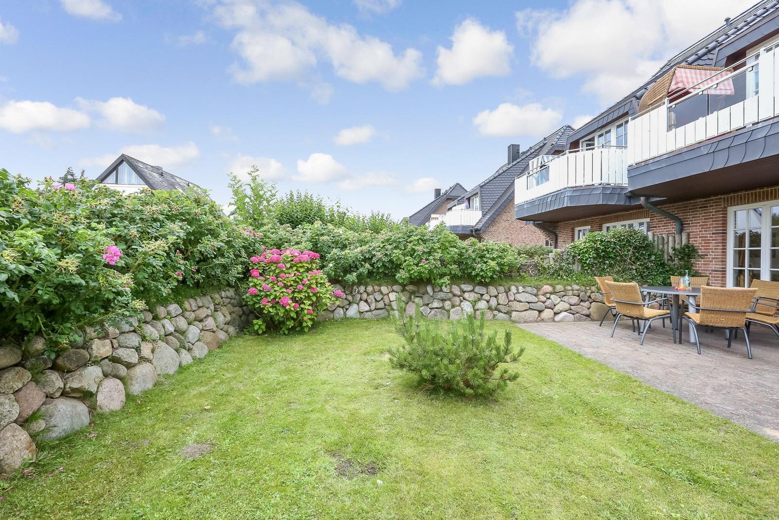 Hinterhof mit Rasen, Steinmauer und Blumen. Terrasse mit Tisch und Stühlen.