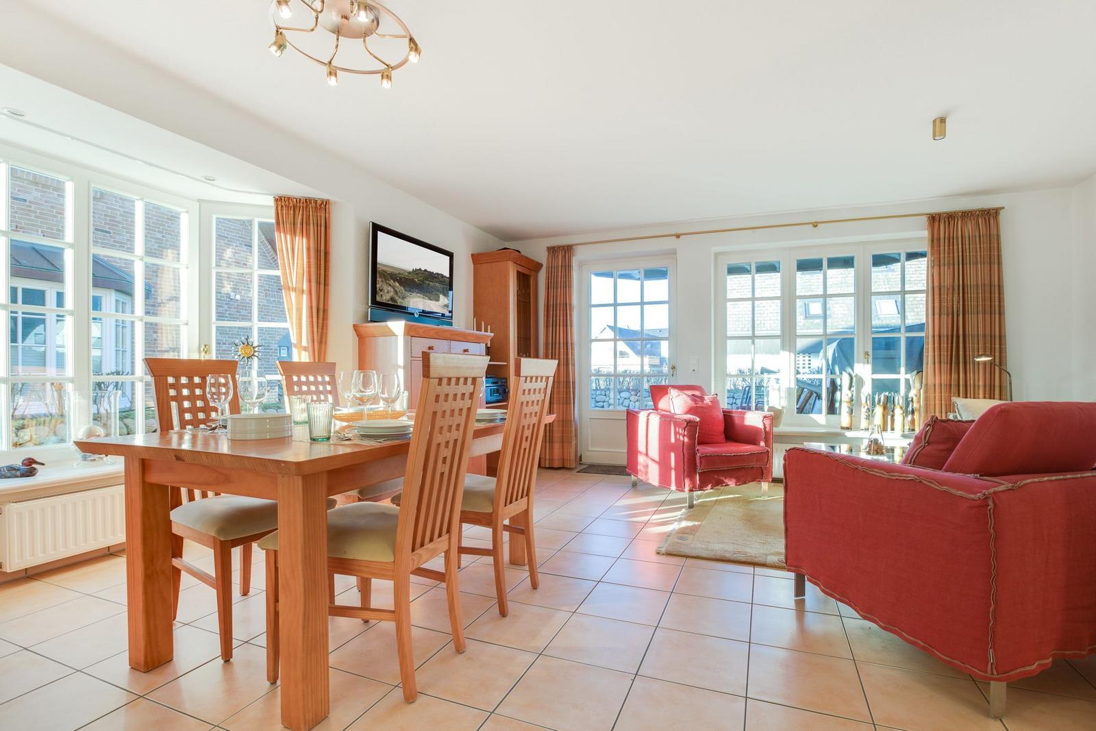 Ein Essbereich mit Holztisch und Stühlen. Wohnbereich mit rotem Sofa und Sessel. Große Fenster und Fliesenboden.
