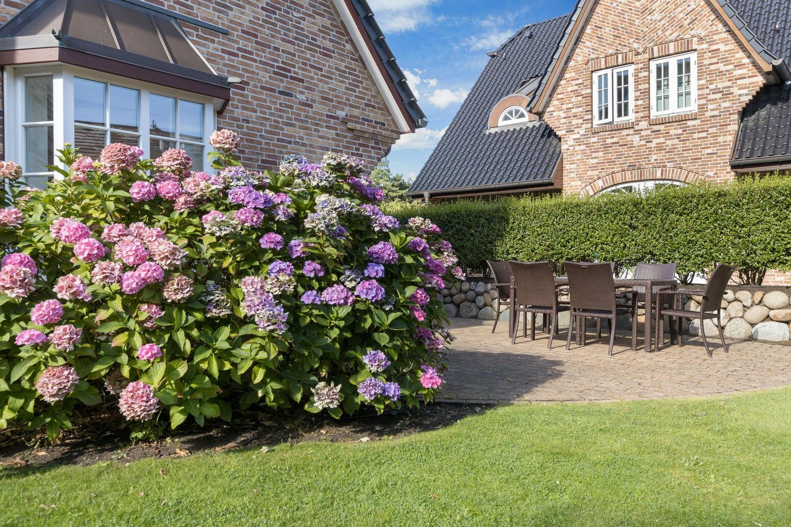 Terrasse mit Sitzgruppe, Blumenrabatten und Grünfläche vor einem Backsteinhaus.